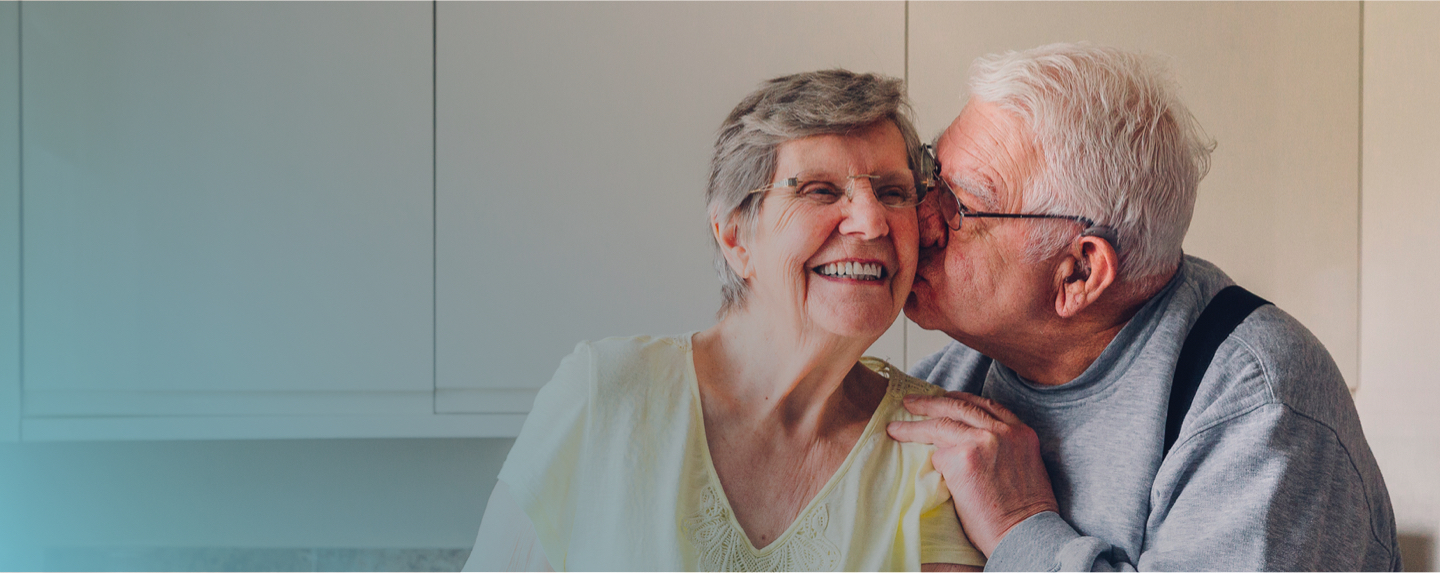 Photograph of an older man with a hearing aid giving his wife a kiss on the cheek