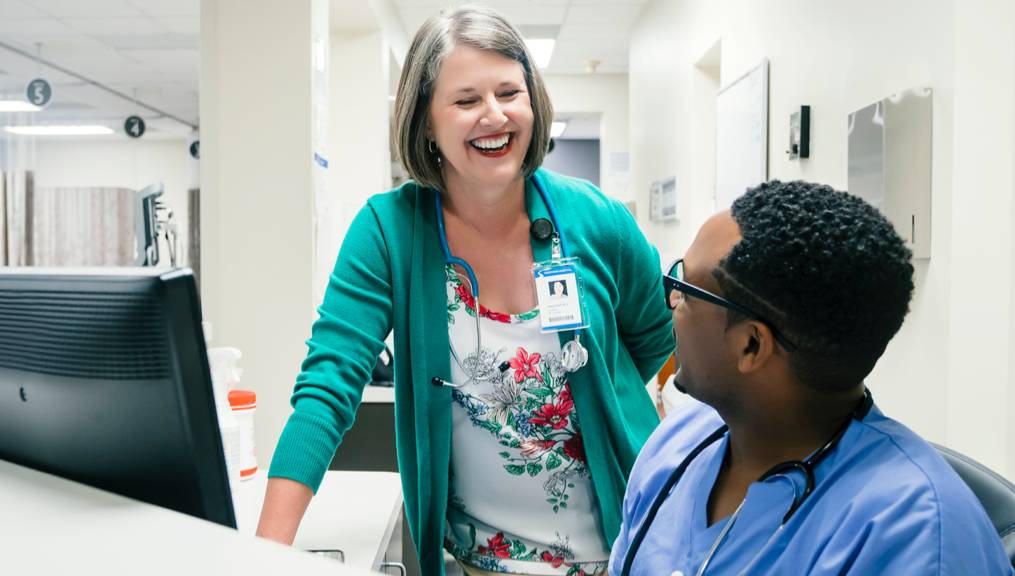 Photograph of two nurses smiling and laughing together in the hallway of a medical office Photograph of two nurses smiling and laughing together in the hallway of a medical office